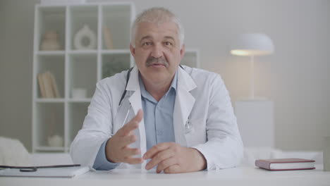 portrait-of-male-therapist-is-speaking-to-camera-sitting-at-table-in-his-office-in-clinic-giving-recommendations